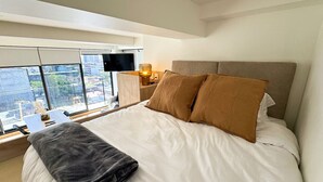 A hotel room with a large bed, a nightstand, a lamp, and a view of the city through large windows. - New apartment Nous 606 (Guadalajara)
