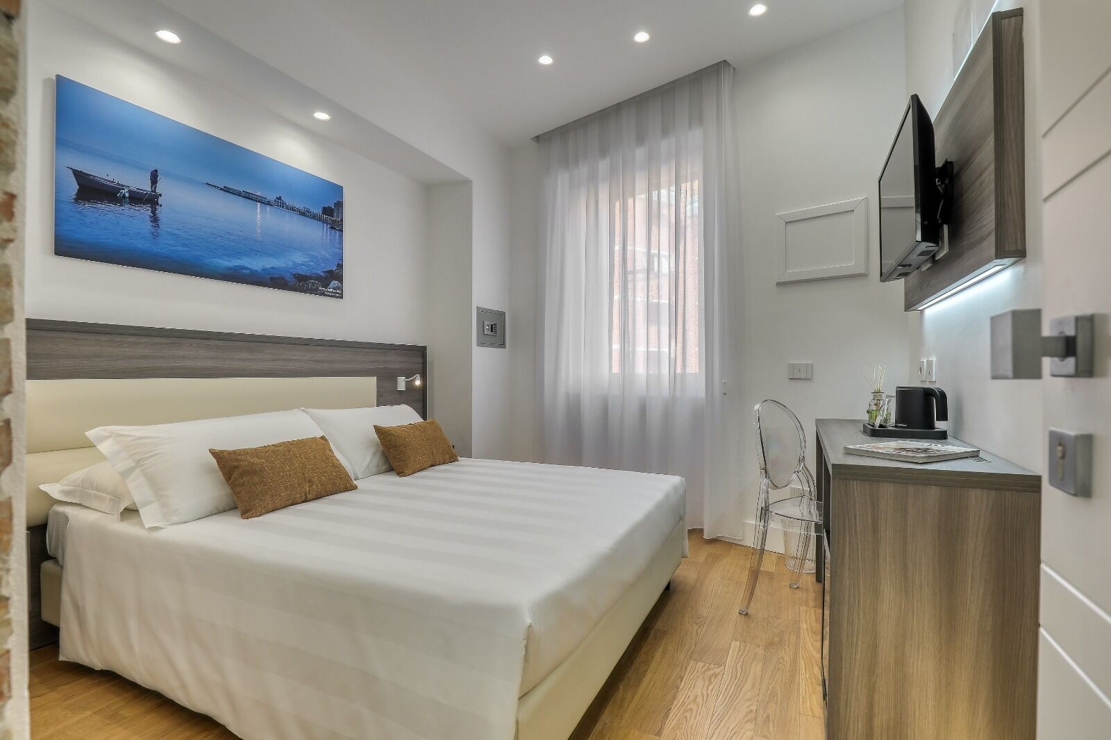 A modern hotel room with a large bed, a flat-screen TV mounted on the wall, a wooden desk with a coffee maker, and a large window with sheer curtains.