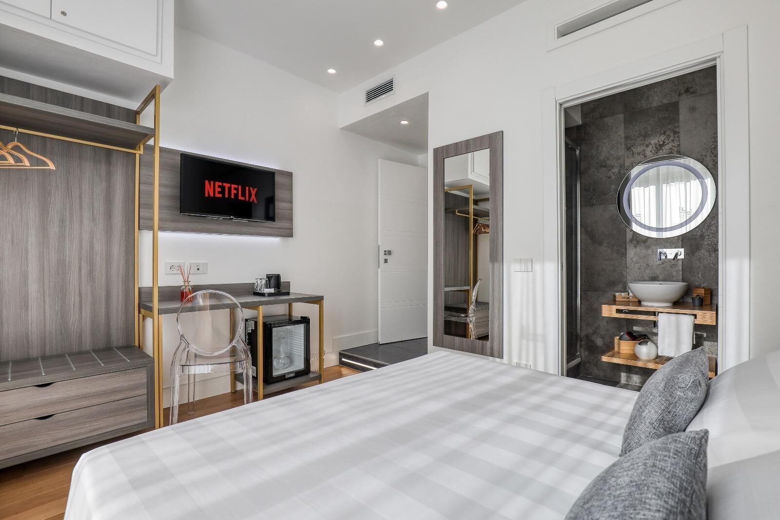 Una habitación de hotel moderna con cama, escritorio con computadora, televisor mostrando 'NETFLIX', armario, baño con espejo y lavamanos, y un espejo redondo.