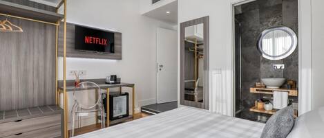 Una habitación de hotel moderna con cama, escritorio con computadora, televisor mostrando 'NETFLIX', armario, baño con espejo y lavamanos, y un espejo redondo.