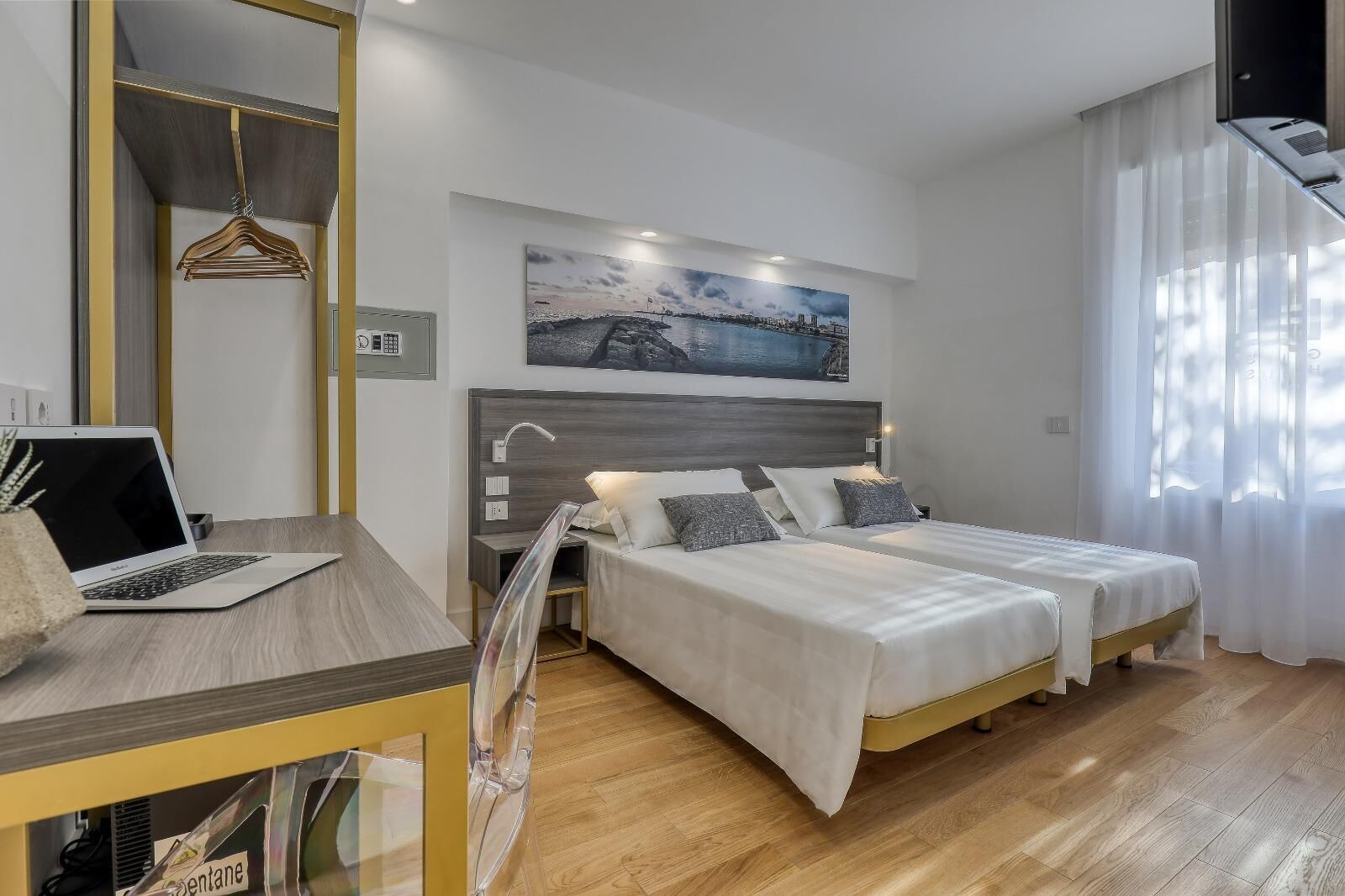 A modern bedroom with a wooden desk, a laptop, two beds with white bedding, a nightstand, a painting, and a large window with sheer curtains.