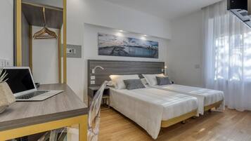 A modern bedroom with a wooden desk, a laptop, two beds with white bedding, a nightstand, a painting, and a large window with sheer curtains.