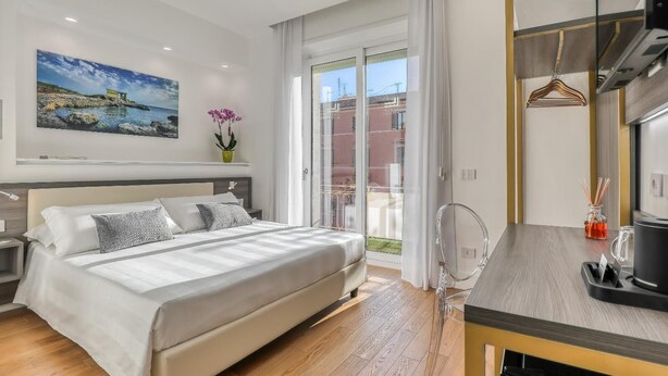 A modern bedroom with a large bed, a desk, and a view of the city through the window. - Gallery Suite (Civitavecchia)
