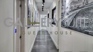 A corridor with a modern design, featuring a long hallway with doors on the left and a wall with framed pictures on the right. The text 'GALLERY SUITE GUEST HOUSE' is prominently displayed. - Gallery Suite (Civitavecchia)