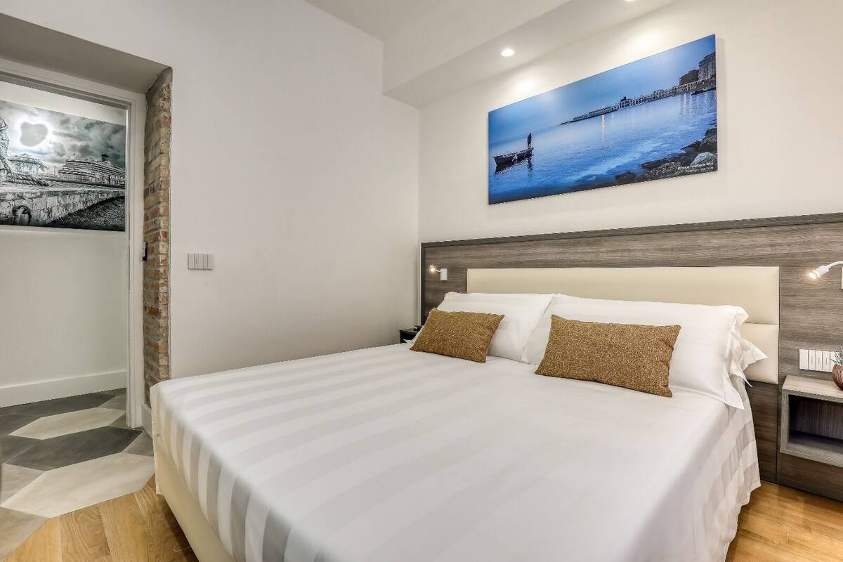 A hotel room with a large bed, a headboard, a nightstand, and a wall-mounted picture of a coastal scene.