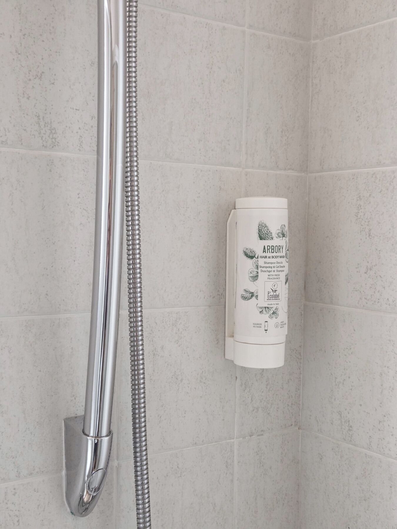 A shower with a chrome hose and a white bottle of Arbory hair and body wash on the wall.