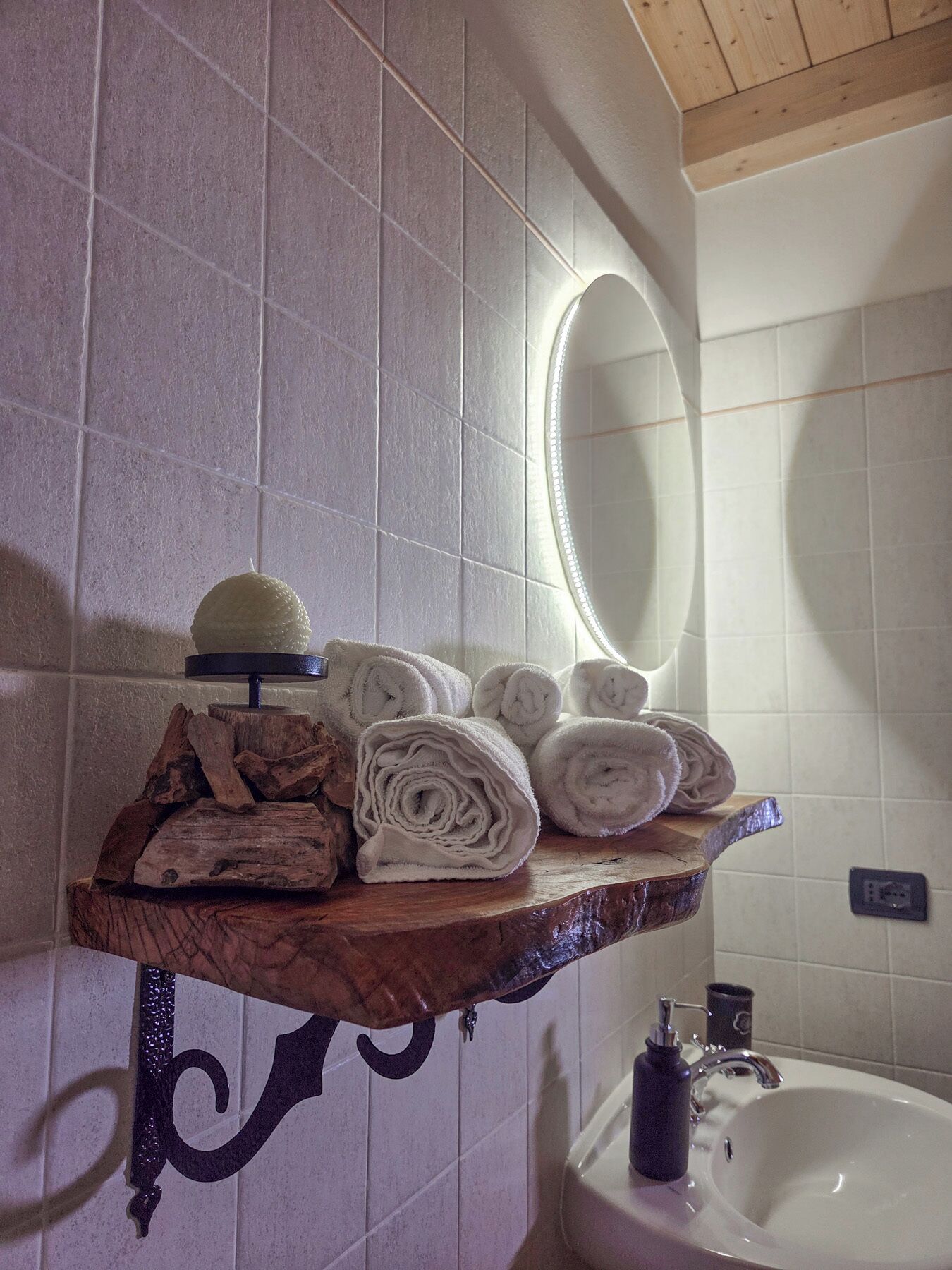 Une salle de bain avec une étagère en bois où sont rangées des serviettes pliées et un chapeau, un lavabo et un miroir.
