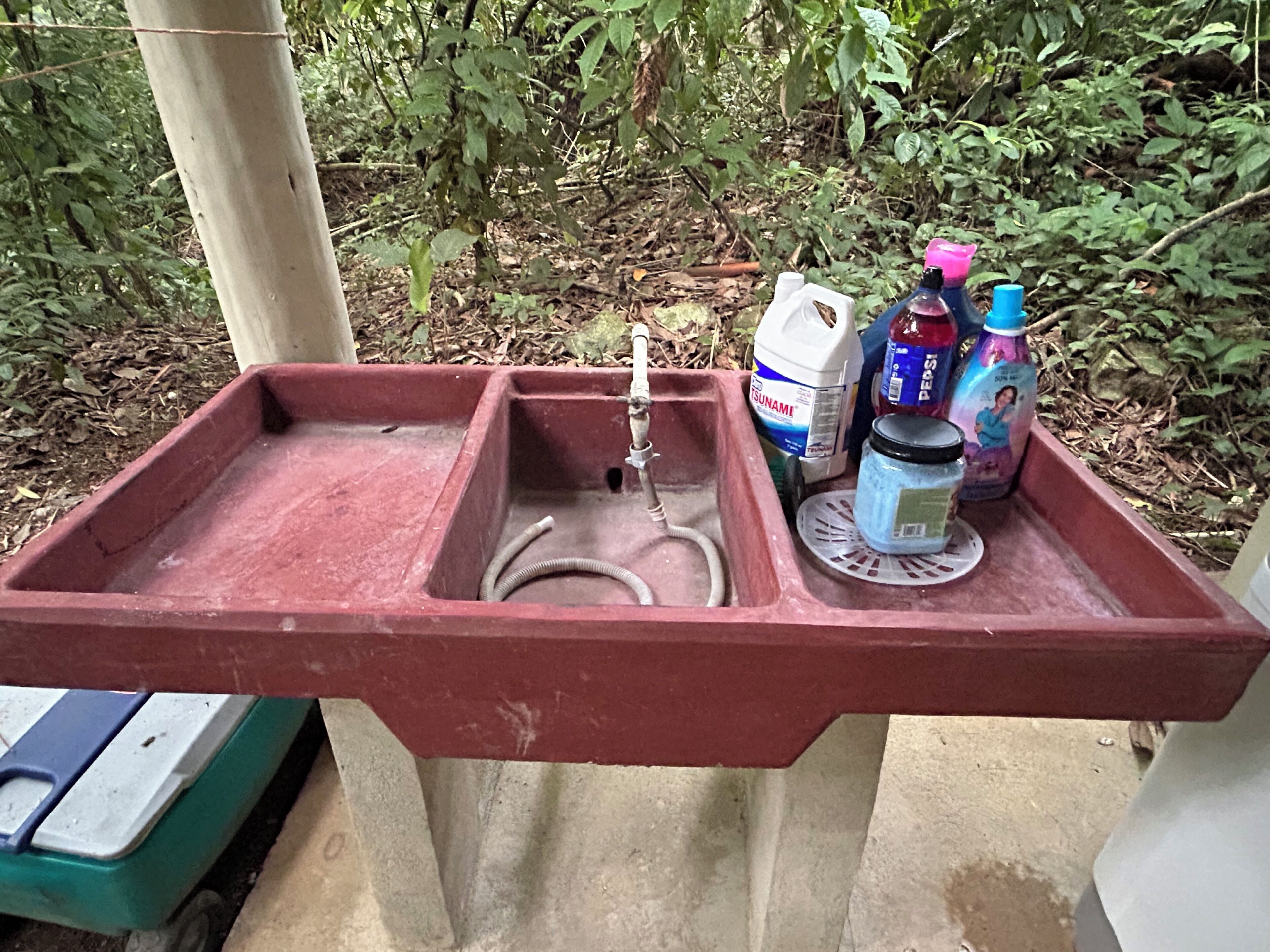 A red metal sink with a hose, a white container labeled 'TSUNAMI', and two bottles of 'PEPSI' and 'Coca-Cola'.