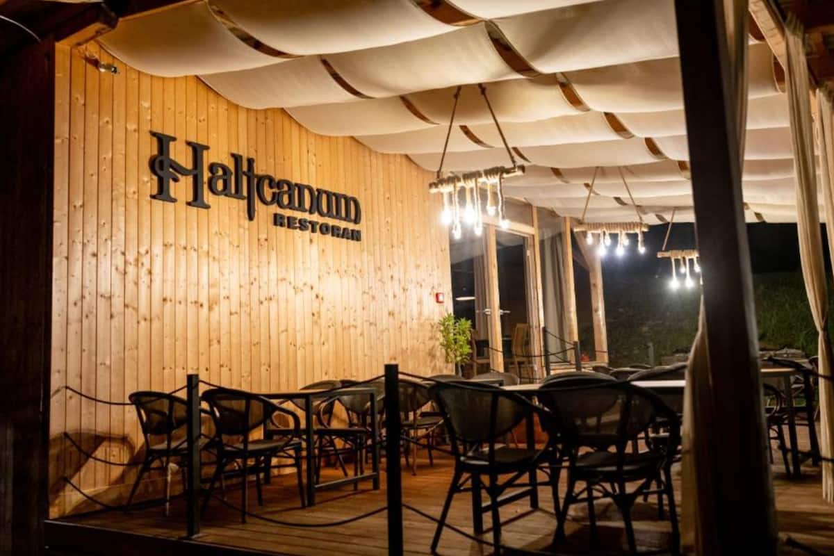 An outdoor seating area with wooden tables and chairs, a sign reading "Halfcannon RESTORAN", and a wooden wall backdrop.