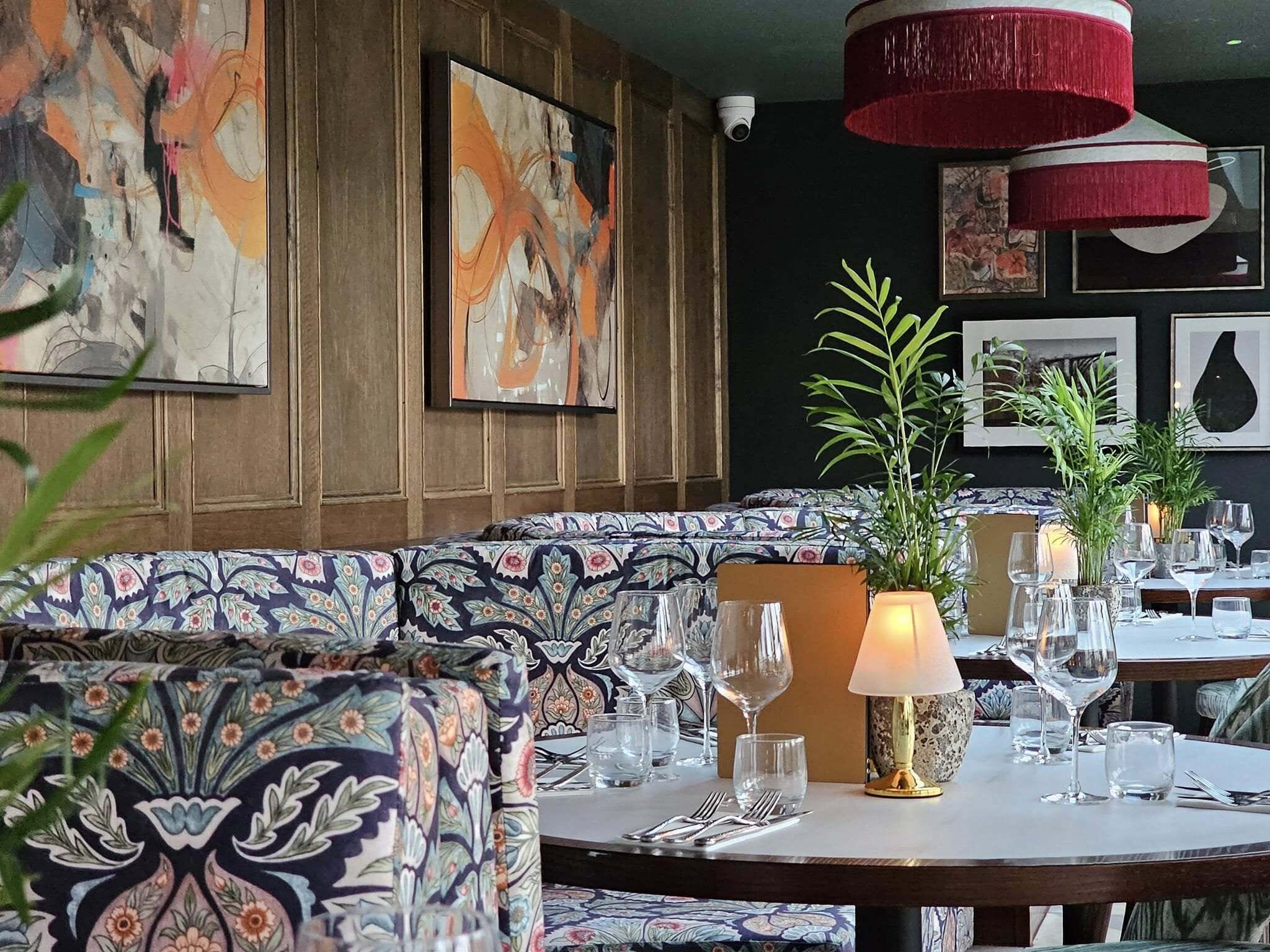 En restaurantindretning med mønstrede siddepladser, et bord dækket med vinglas og billedkunst på væggene.