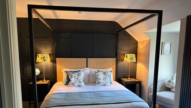 A four-poster bed with a dark headboard, two bedside lamps, and a patterned ottoman. - The Sutherland (Telford)