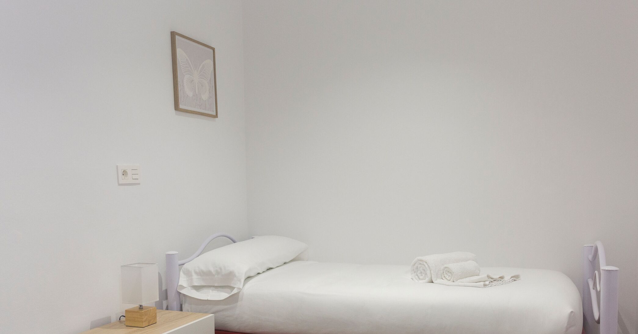 Una habitación pequeña y limpia con una cama individual, una mesita de noche y una lámpara en la pared.
