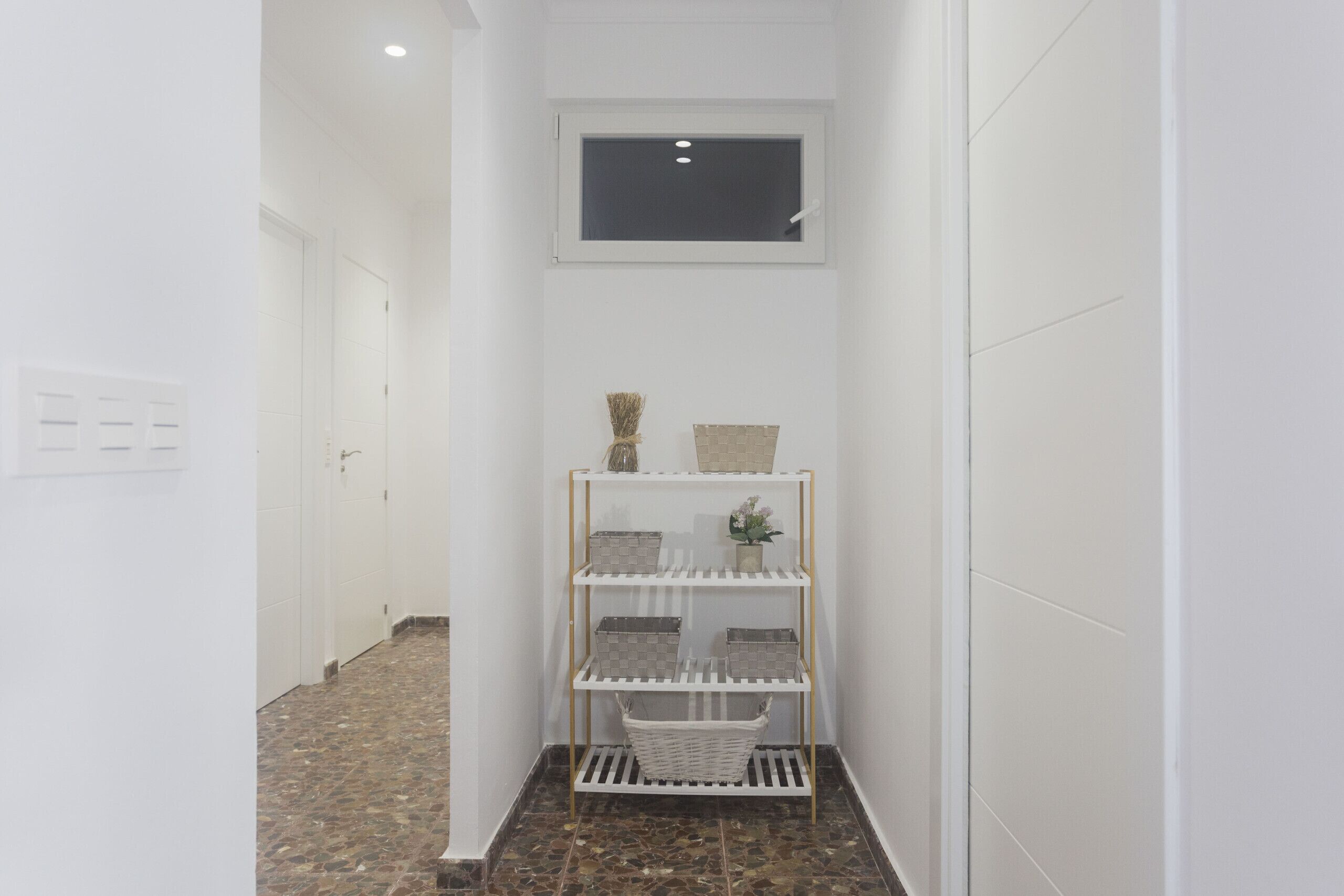 A narrow hallway with a white tiled floor, a white wall with a window, and a white shelf with baskets and a vase.