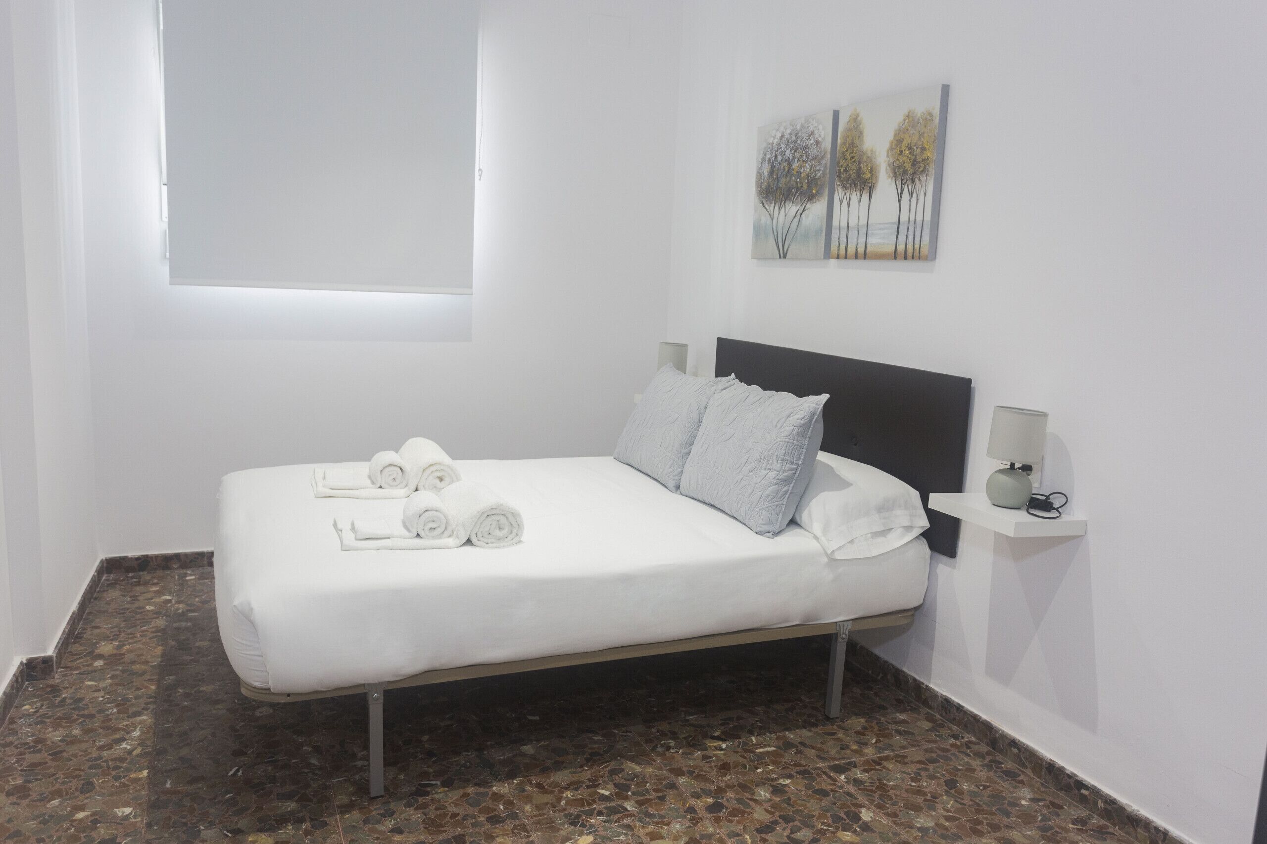A neatly made bed with white linens and towels, a headboard, a wall-mounted lamp, and a painting of trees.