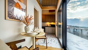 Un coin repas avec une table ronde, des chaises et un vase contenant des plantes séchées. On aperçoit une cuisine à l’arrière-plan, avec des meubles en bois et une fenêtre donnant sur un paysage enneigé.