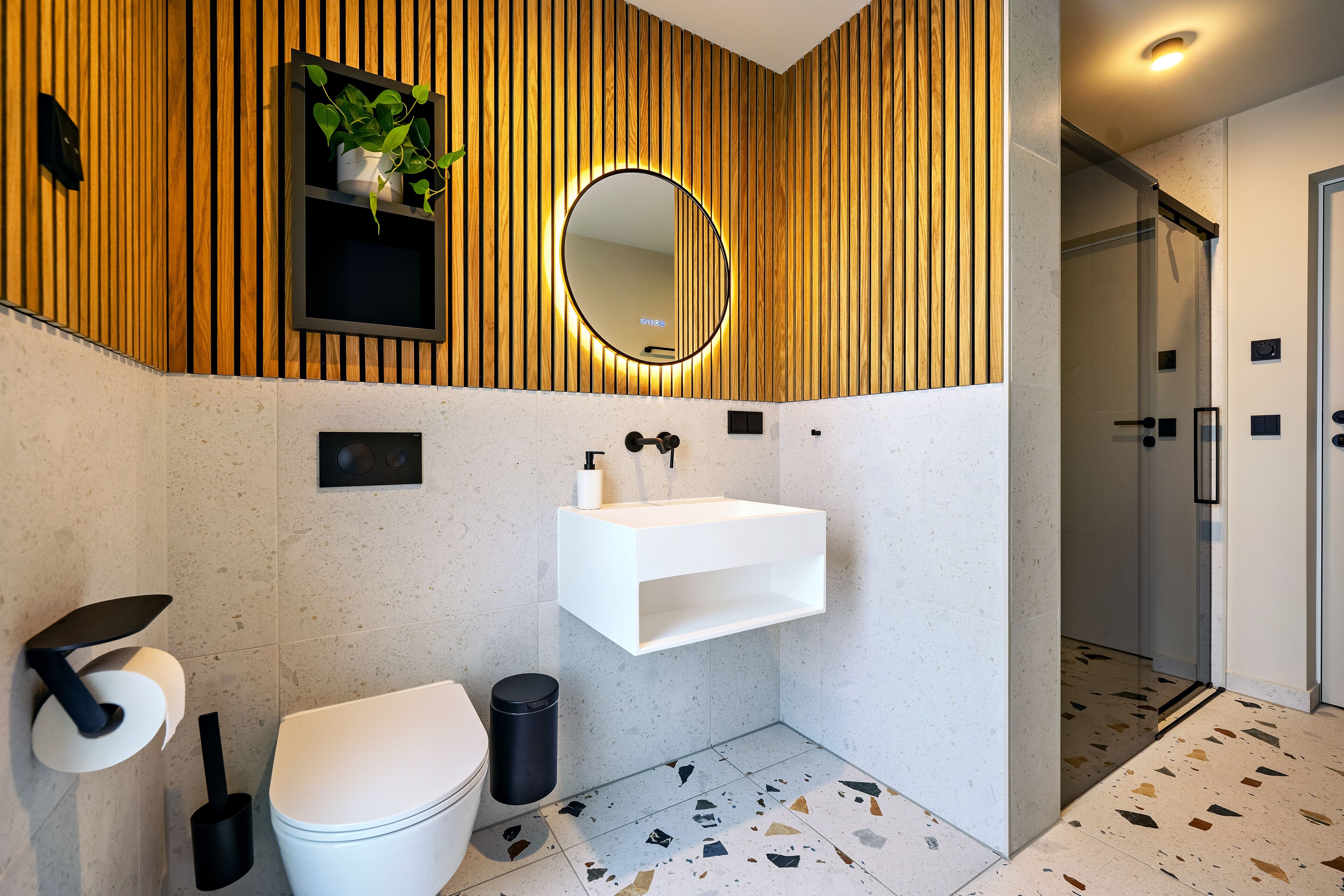 Ein modernes Badezimmer mit einer Wand aus Holzpaneelen, einem runden Spiegel, einem weißen Waschbecken und einer Toilette.