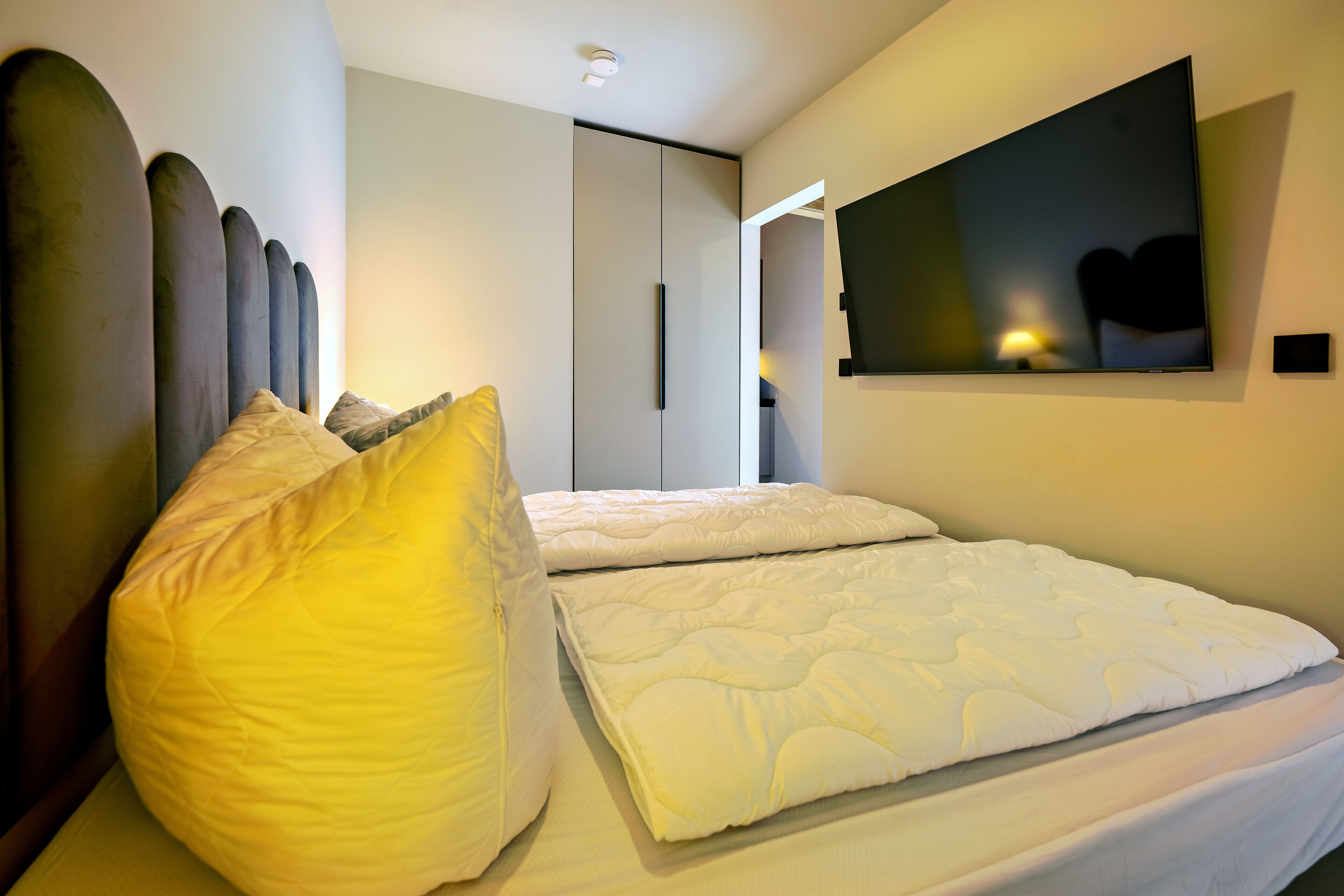 A modern bedroom with a large bed, a flat-screen TV mounted on the wall, and a wardrobe with a sliding door.