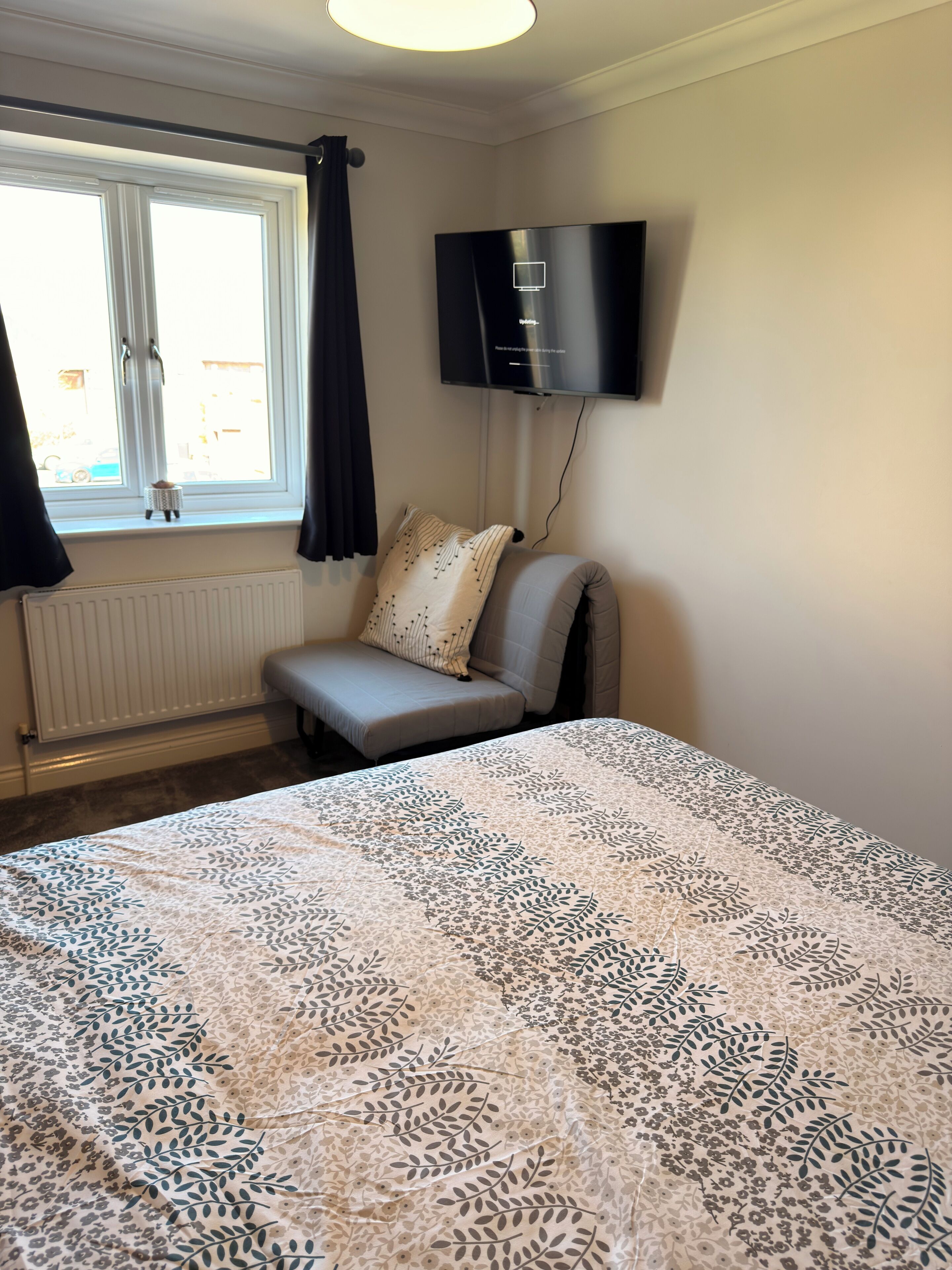 A bedroom with a bed, a window with curtains, a radiator, a TV mounted on the wall, and a small table with a lamp.