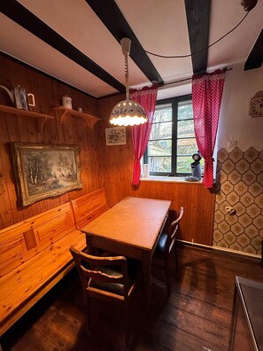 A wooden interior with a table and benches, a window with curtains, and a wall-mounted clock.