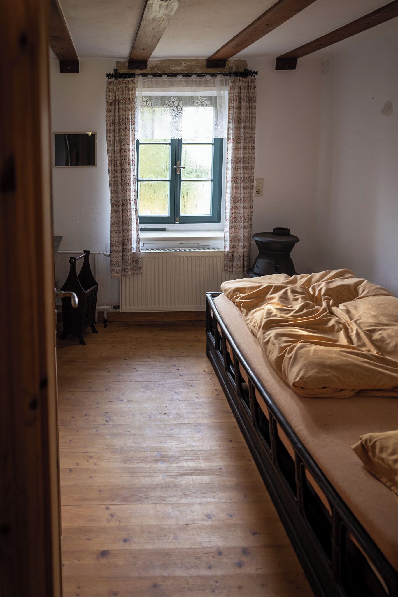 A narrow room with a wooden floor, a bed with a beige blanket, a window with floral curtains, a radiator, and a small table with a lamp.