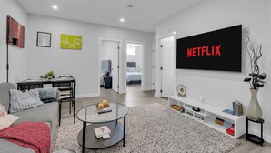 A modern living room with a flat-screen TV displaying "NETFLIX," a gray sofa, a coffee table, and a dining area in the background.