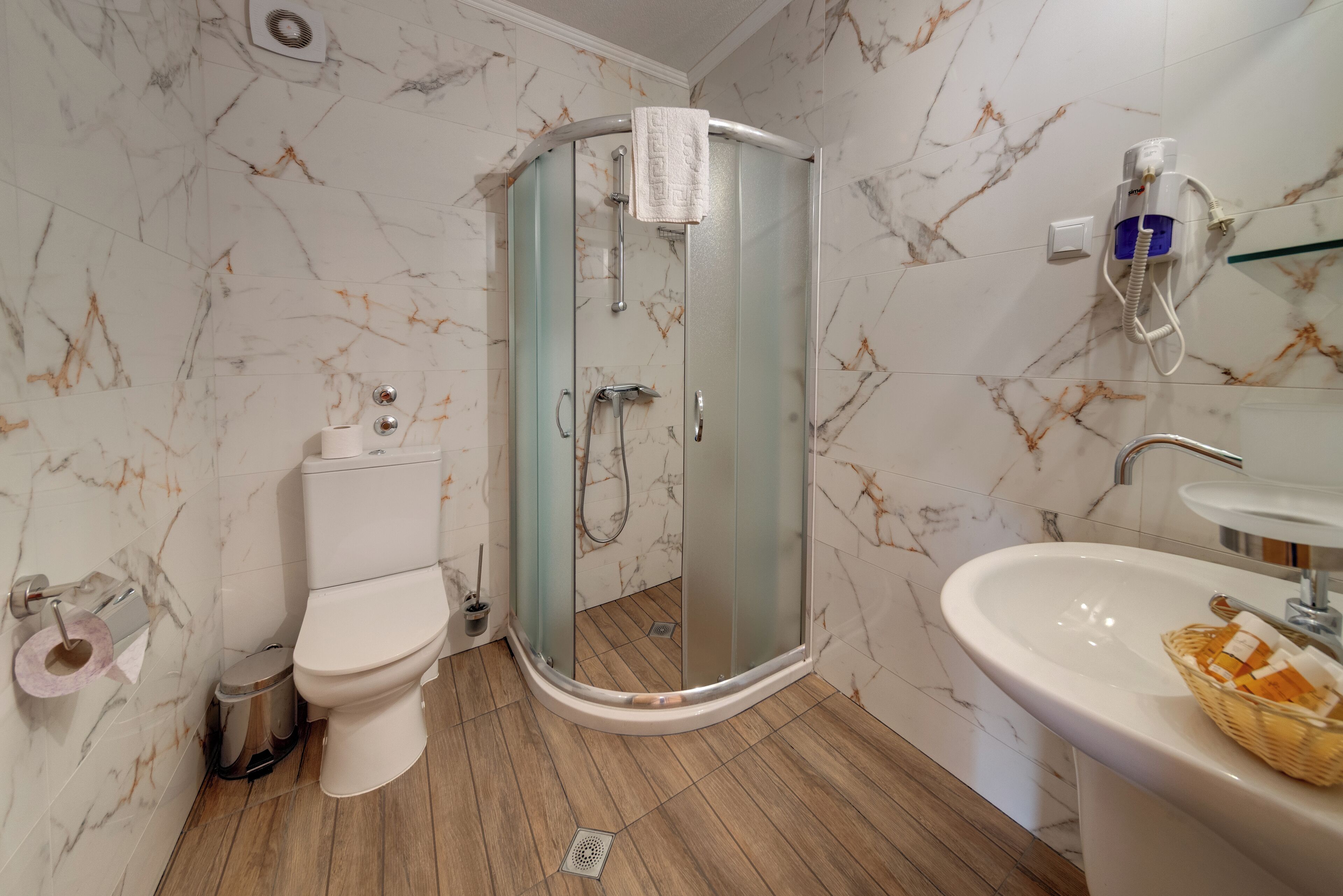 Un baño con inodoro, ducha con mampara de vidrio, lavamanos y paredes con diseño de mármol.