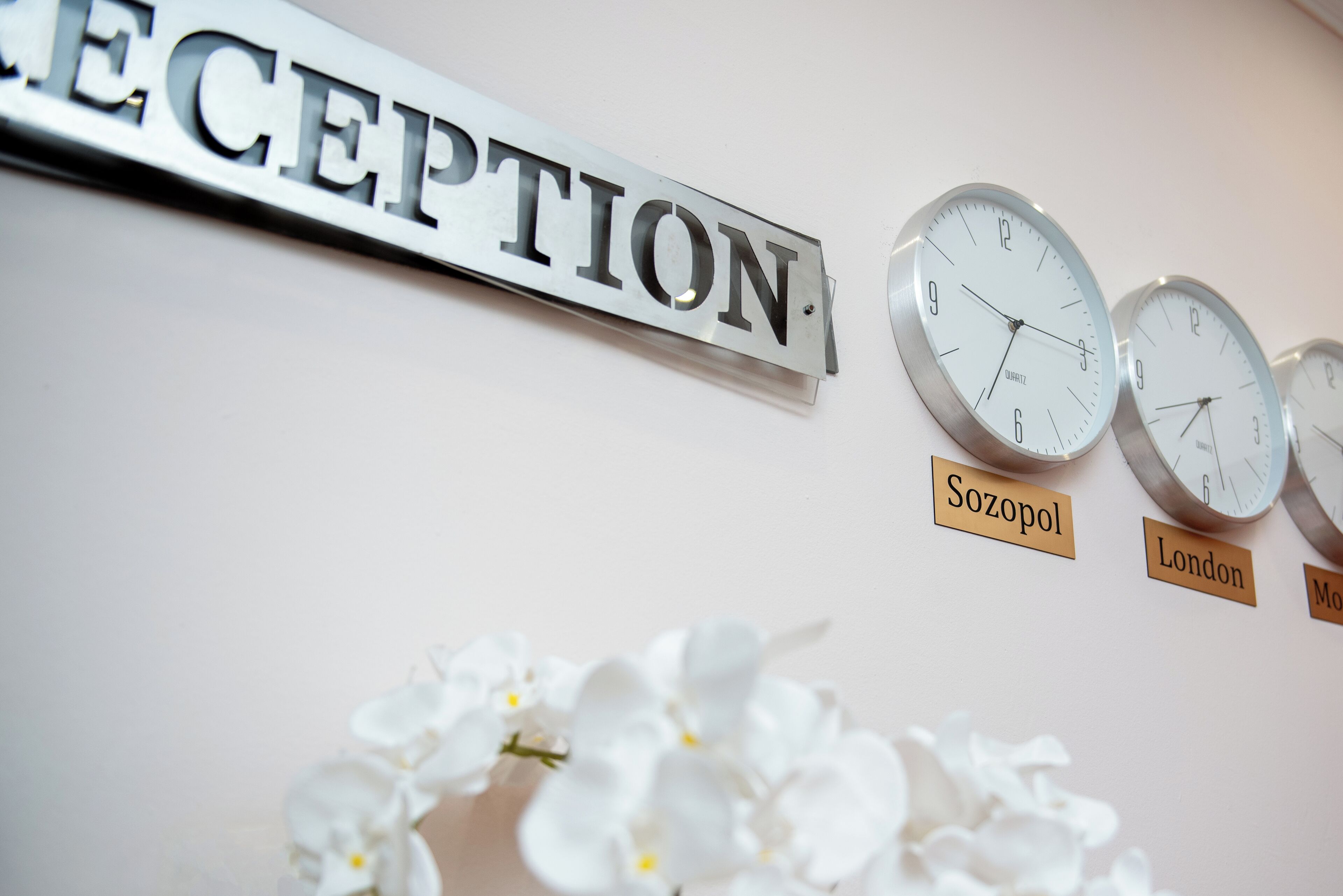 Un'area reception con un cartello che indica 'RECEPTION', orologi etichettati 'Sozopol' e 'Londra', e fiori bianchi.