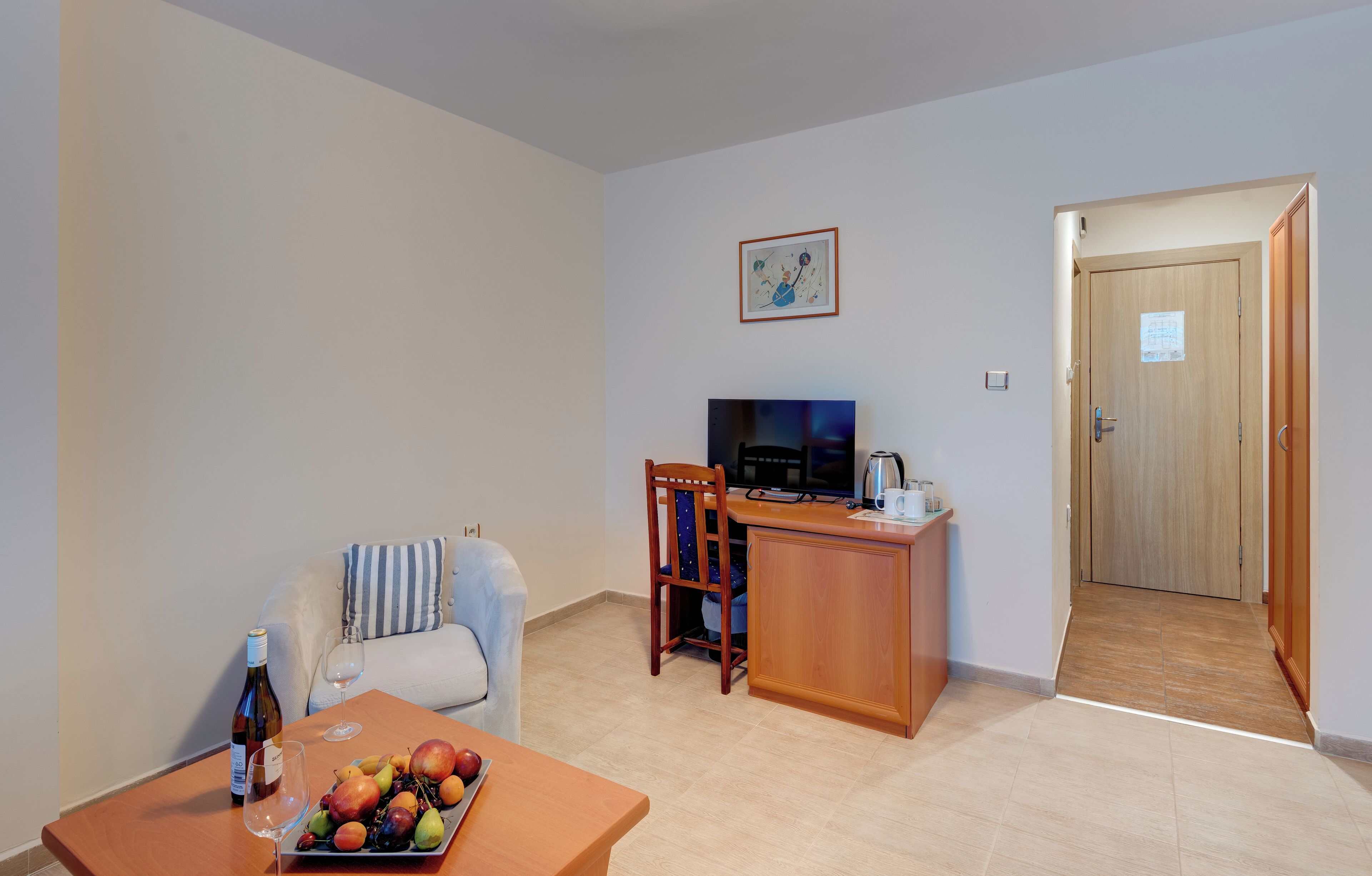 A living room with a sofa, a dining table with fruit and a bottle, a TV, and a wooden door.