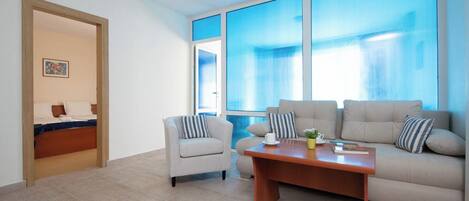 Una sala de estar moderna con sofá, sillón y mesa de centro. Hay una cama en la habitación contigua, y ventanas amplias que dejan entrar luz natural.