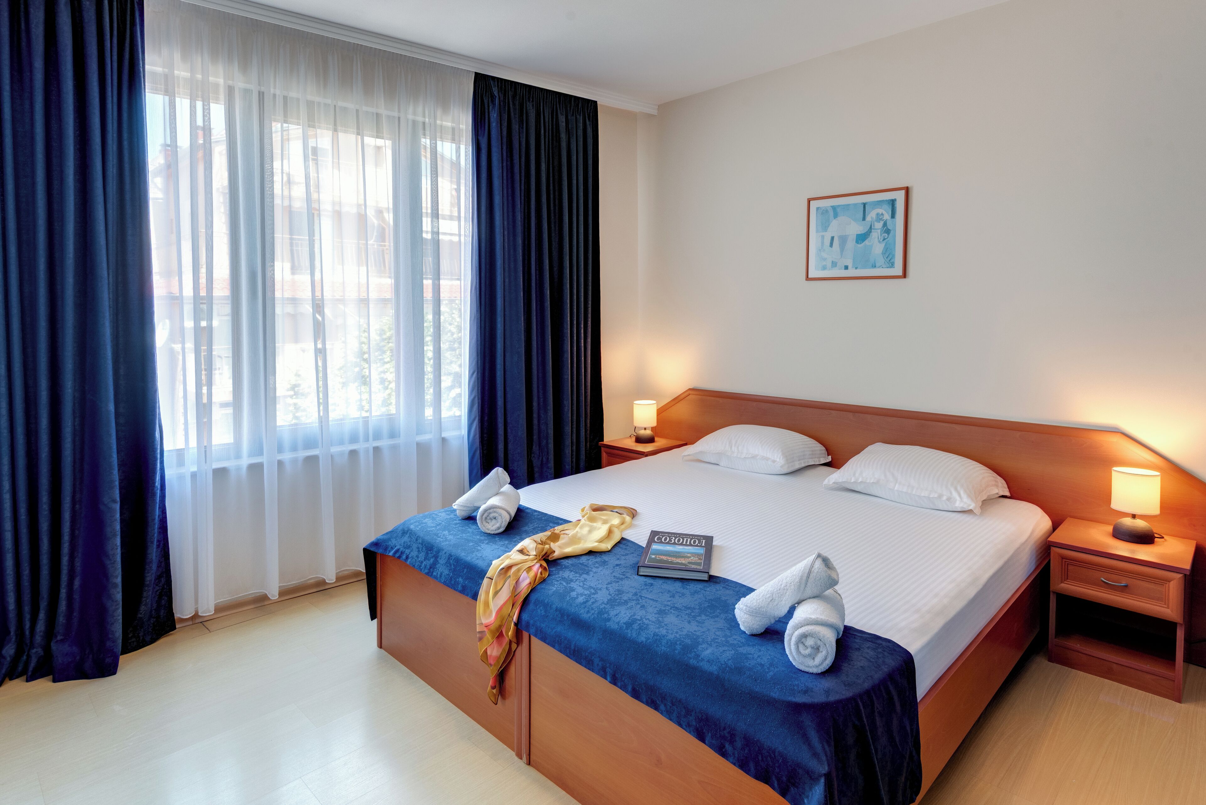A neatly made bed with white linens, a blue bedspread, and a book titled "Cosmos 1". There are two bedside tables with lamps, a framed picture on the wall, and a window with sheer curtains.