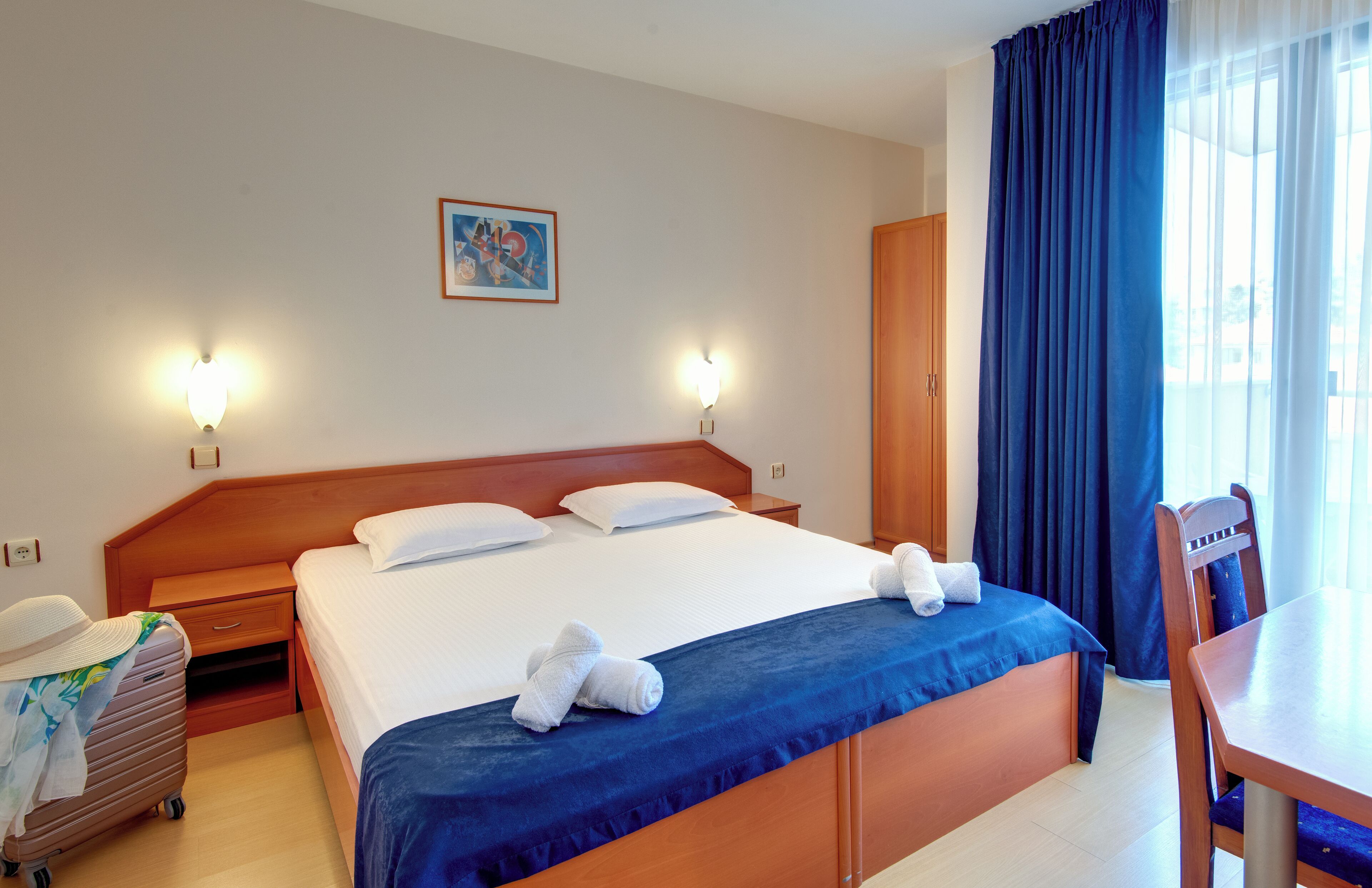 A hotel room with a large bed, two bedside tables, a wooden wardrobe, a dining table, and a chair.