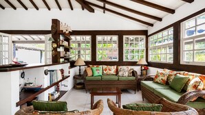 Una habitación con techos altos, vigas de madera, un conjunto de sofá verde, muebles de mimbre y ventanas amplias con vistas a la vegetación.