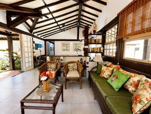 A room with a high ceiling, wooden beams, a green sofa with floral cushions, wicker chairs, and a glass coffee table with a vase of flowers. - Bz39 Casa 150m da praia piscina priv (Buzios)