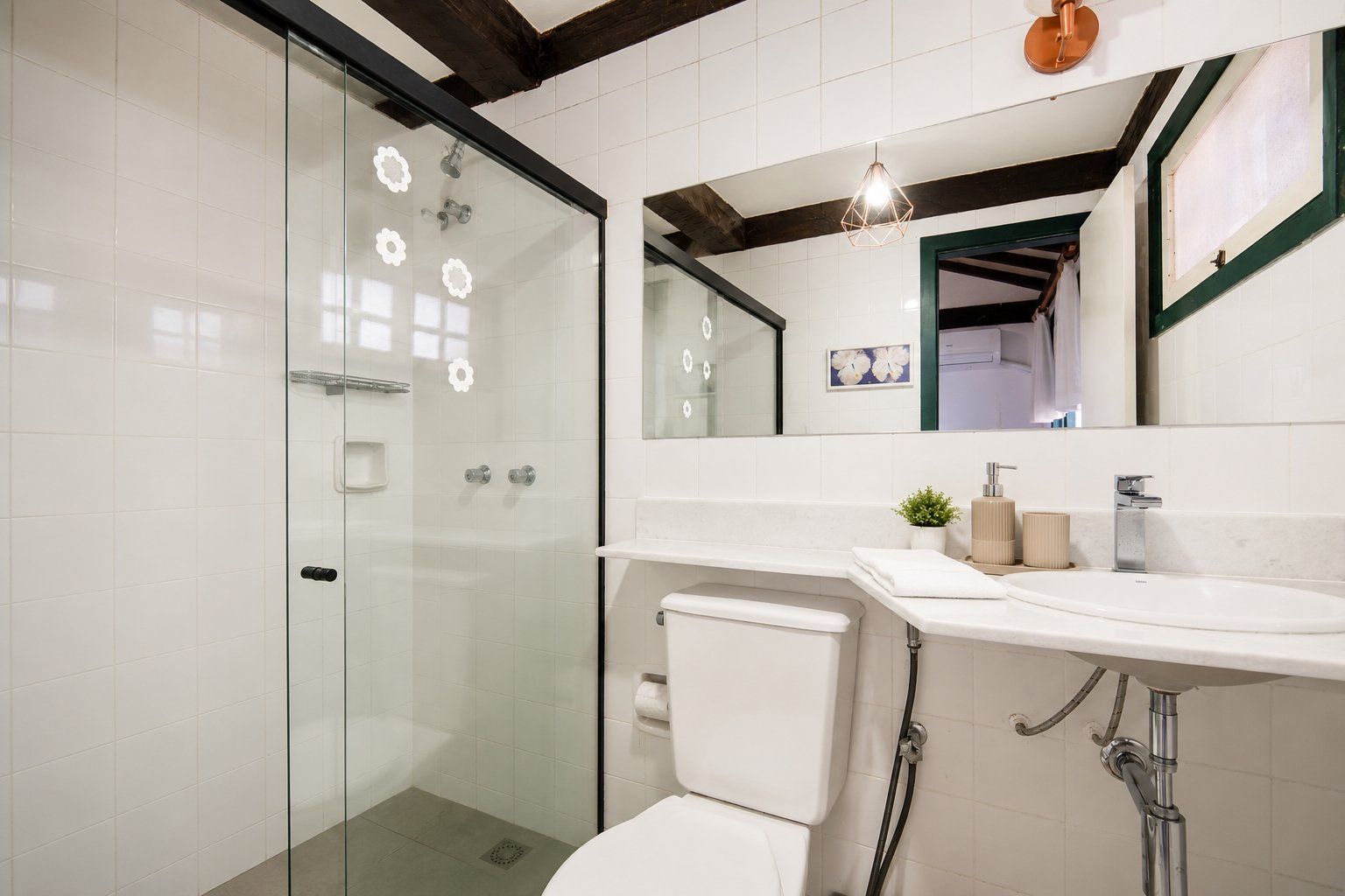 Et moderne badeværelse med en brusekabine i glas, et hvidt toilet, en vask med et spejl over, og en træbjælke i loftet.