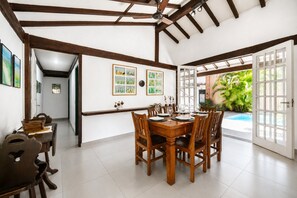 Un coin repas avec une table et des chaises en bois, un ventilateur de plafond et une vue sur une piscine, visible à travers une porte ouverte.