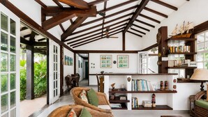 Amplia habitación con techo alto, vigas de madera a la vista, zona de comedor y una estantería llena de libros y objetos decorativos.