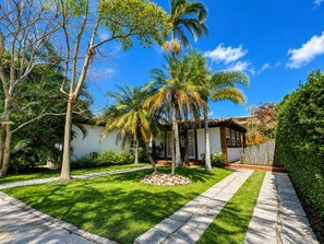 Una propiedad residencial con un césped bien cuidado, un camino de piedra y una variedad de árboles, incluyendo palmeras.