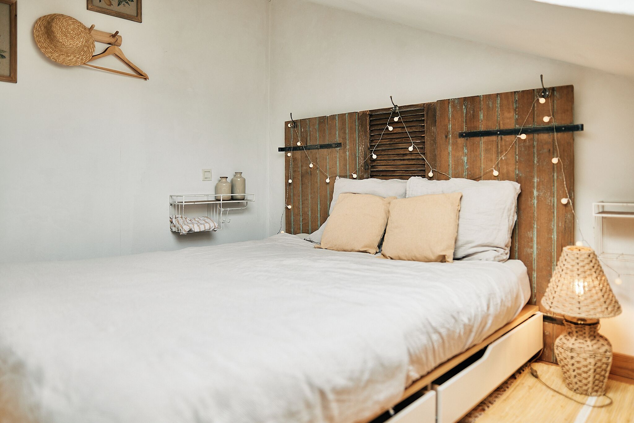A bedroom with a wooden headboard, a bed with white bedding, a bedside lamp, and a wall-mounted shelf with decorative items.