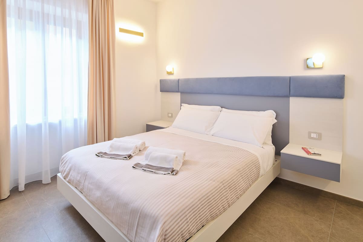 Un lit bien fait, avec du linge de lit blanc et deux serviettes, une table de chevet avec un livre, et un luminaire fixé au mur.