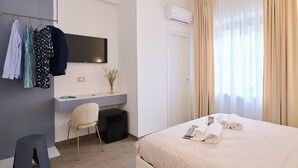 A hotel room with a bed, a desk, a chair, a small stool, a TV, and a wardrobe. - San Nicola Rooms Siracusa (Syracuse)
