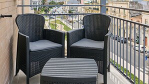 A balcony with two wicker chairs and a matching ottoman, overlooking a cityscape with buildings and a road. - San Nicola Rooms Siracusa (Syracuse)