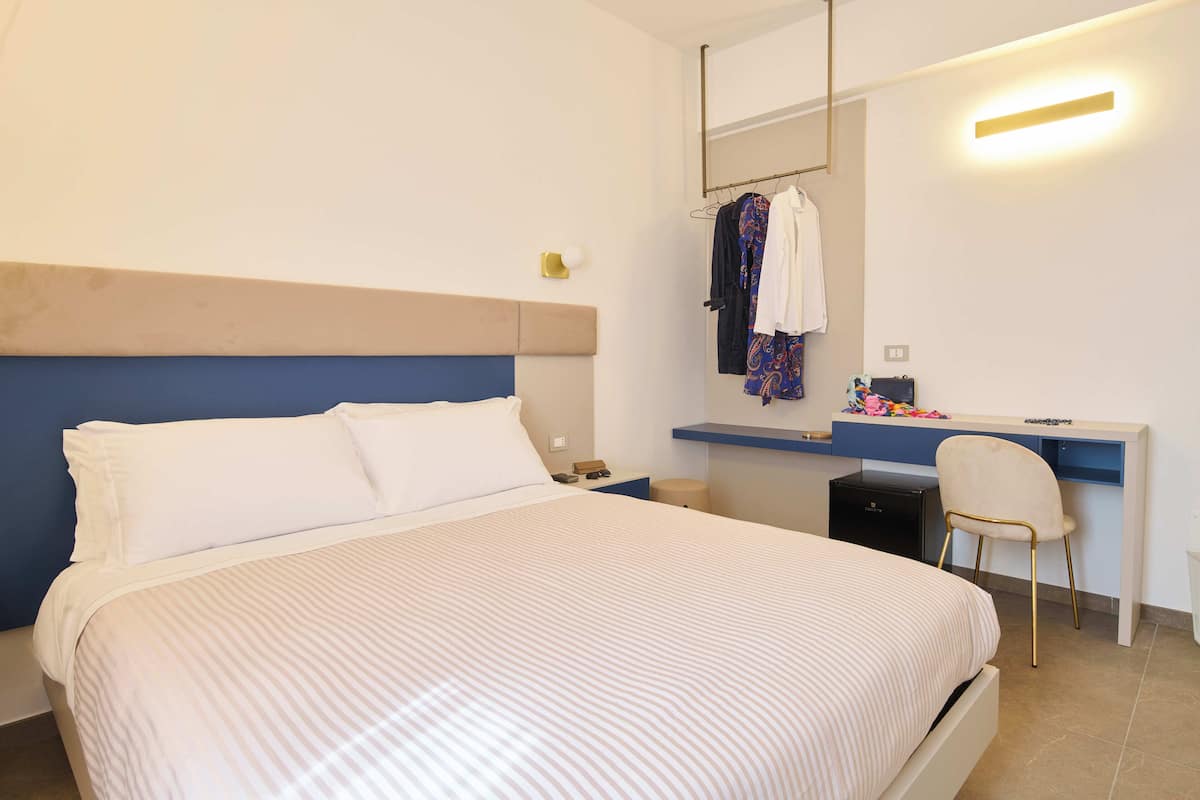 Un lit bien fait, recouvert d’une couvre-lit à rayures, une table de chevet avec une lampe, une chaise et un bureau avec un miroir et des vêtements suspendus au-dessus.