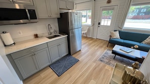 A modern kitchen with grey cabinets, a stainless steel refrigerator, and a microwave. There is a wooden floor, a blue sofa, and a window with curtains. - New - The Skyline Flat (Front Royal)