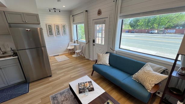 A modern living room with a blue sofa, a coffee table, and a kitchen area with a stainless steel refrigerator. - New - The Skyline Flat (Front Royal)