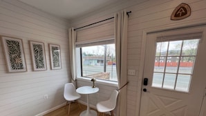 A room with white paneled walls, a wooden floor, a small round table with two white chairs, a window with curtains, and a door with a coat of arms. - New - The Skyline Flat (Front Royal)