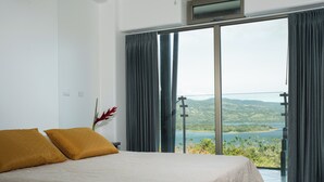 Een slaapkamer met een groot bed, twee kussens, een nachtkastje en uitzicht op een meer en heuvels door een schuifdeur.