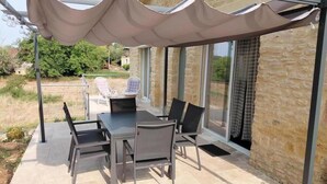 A covered patio with a table and chairs, a lounge chair, and a glass door leading to a garden.