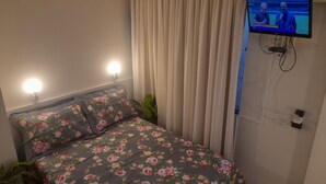 A bedroom with a floral bedspread, two bedside lamps, a TV mounted on the wall, and a curtain. - Air-conditioned studio, wifi in the center of Piratuba SC (Piratuba)