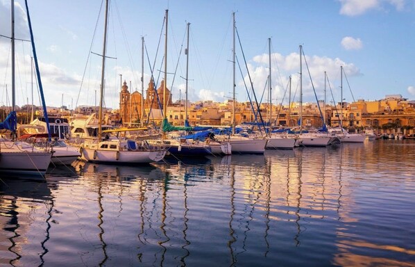 A marina with multiple boats docked, a prominent building with a bell tower in the background, and calm water reflecting the boats.