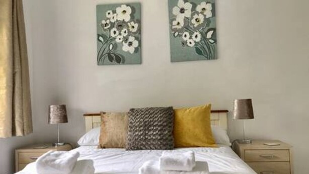 A neatly made bed with white linens, a gray and yellow pillow, and a wall-mounted lamp beside it. - The Woodfarm Lodge - 3 Bedroom House With Parking (Oxford)